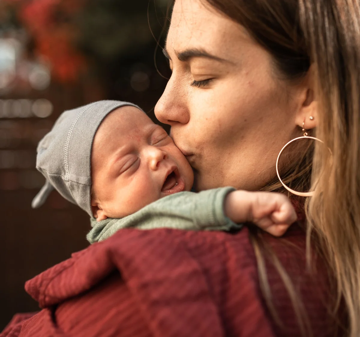 Mamá dándole un beso en la mejilla a su bebé recién nacido, con gorrito gris, bajo los árboles de otoño